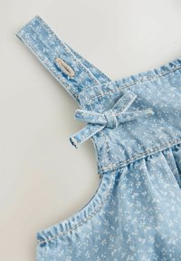 Bretelle de salopette en denim bleu clair avec boutonnière cousue et nœud noué, montrant une partie du tissu froncé en dessous.
