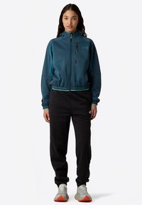 Tyrkysová bunda na zip s černými doplňky a malou kapsou na zip, doplněná černými tepláky s logem. Tenisky mají oranžové podrážky.