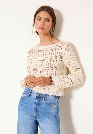 Woman with brown hair wearing a cream-colored crochet sweater and blue jeans, standing against a white wall with hands clasped.