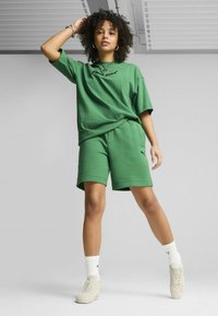 Camiseta de algodón verde con texto bordado en negro y pantalones cortos verdes a juego. El modelo lleva calcetines blancos y zapatillas crema, con cabello rizado y texturizado.