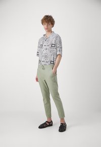 Kurzarmhemd mit schwarz-weißem Muster, kombiniert mit hellgrünen Hosen und schwarzen Sandalen mit Schnallen.