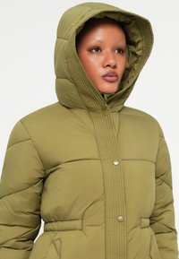 Casaco puffer verde-oliva com capuz, apresentando painéis acolchoados, fecho de botão e cintura ajustada para uma silhueta ajustada.