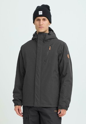 Noor mees musta talvejaki ja lukuga rinnatasku ning valge logoga musta mütsi ja taustal seismas.