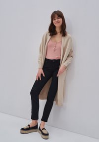 Bež dug kardigan preko ružičaste majice s dugim rukavima i dugmadi, uparen s crnim uskim trapericama i crnim espadrilama s bež detaljima.