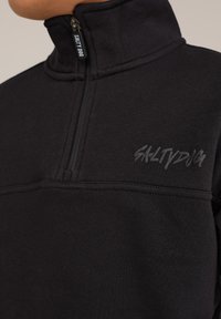 Melnā sporta jaku ar rāvējslēdzēja apkakli, izgatavota no struktūras auduma. Tam ir daļējs rāvējslēdzējs un kontrastējoša melna izšuvuma "SALTY DOG" logo uz krūškurvja.