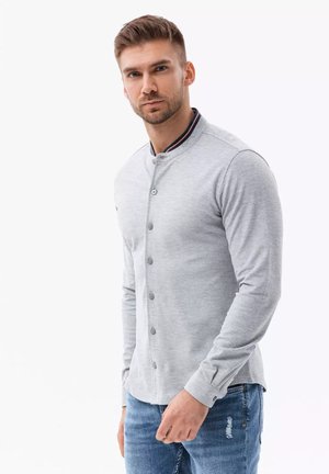 Grijze blouse met lange mouwen en een mandarinnekraag, vooraan knopen en contrasterende marineblauwe en rode accentueringen. Gepaard met lichtblauwe distressed jeans.