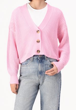 Kvinde iført en pink, chunky strikket cropped cardigan med tre knapper over en hvid top, kombineret med lyseblå jeans med rhinstenmønstre.