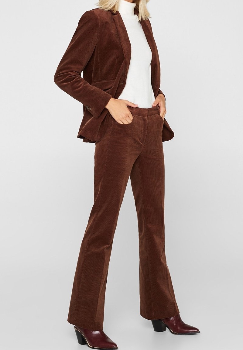 Completo giacca e pantaloni in corduroy marrone, slim-fit, texture a coste, con un dolcevita bianco sotto, abbinato a stivali con tacco bordeaux.
