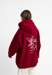 Bordeaux oversized hoodie met een witte lijntekening van een gezicht en bladeren op de achterkant. Voorzien van een grote capuchon en geribde manchetten.