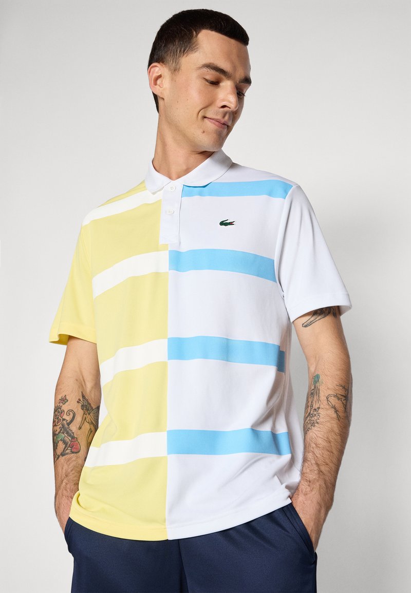 Polo shirt avec des rayures horizontales jaunes, blanches et bleu clair, dotée d'un col à boutons et d'un petit logo vert sur la poitrine.