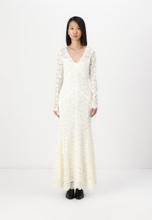 Long-sleeve white lace dress with a scalloped neckline, fitted bodice, and flared skirt; intricate floral patterns, worn with black shoes.