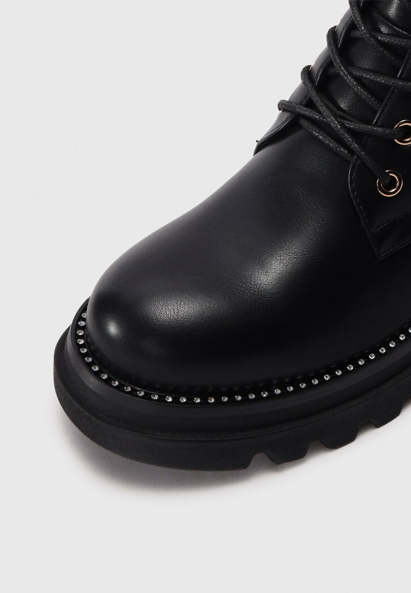 Zwarte leren enkelboot met een ronde neus, zwarte rubberen zool en decoratieve studaccenten langs de rand. Vetersluiting.