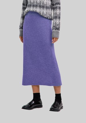 Person trägt einen violetten gestrickten Midirock, gemusterten grauen Pullover, schwarze Socken und schwarze glänzende Loafers und steht vor einem grauen Hintergrund.