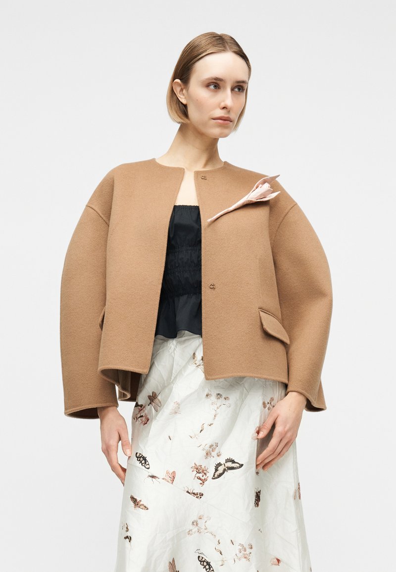 Woman with short blonde hair wearing a camel-colored open jacket with a pink flower pin, black top, and white skirt with butterfly print.