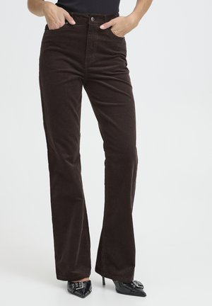 Vrouw die een donkerbruine ribfluwelen hoge taille broek draagt met zwarte pumps met puntige neus, handen in de voorzakken, staand tegen een witte achtergrond.
