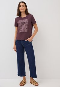 T-shirt à manches courtes de couleur bordeaux foncé avec un motif de feuilles ; associé à un jean bleu à jambes larges et des sandales tissées marron. Fond uni.