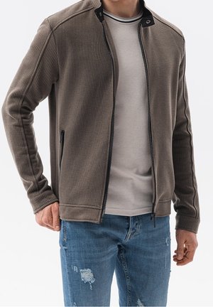 Man i en strukturerad brun zip-up jacka, ljusgrå skjorta och blå slitna denimjeans mot en enfärgad bakgrund.
