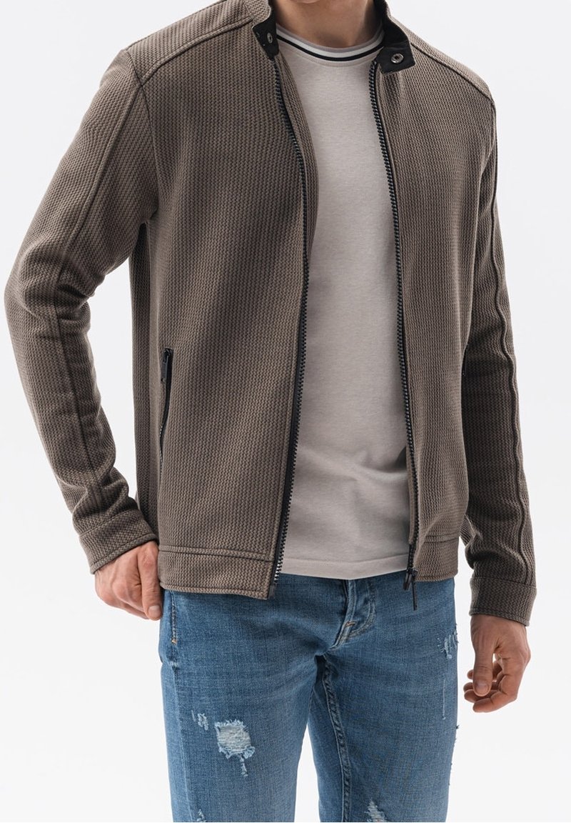 Mężczyzna ubrany w brązową kurtkę z teksturowanym zamkiem, jasnoszary T-shirt i niebieskie zniszczone dżinsy na jednolitym tle.