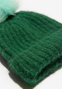 Zelená pletená čepice vyrobená z měkké vlny, má texturovaný žebrovaný design a nahoře světle modrou umělou kožušinu v podobě pom-pomu.