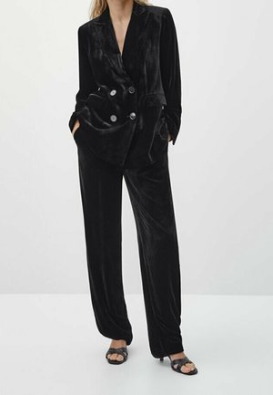 Femme portant un blazer croisé en velours noir avec un pantalon large assorti et des sandales à talons ouvertes noires, les mains dans les poches.