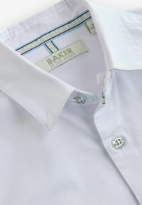 Bílá košile s límečkem a knoflíkovým zapínáním, která má jemnou texturu, kontrastní modré vnitřní švy a detaily stříbrného knoflíku s logem.