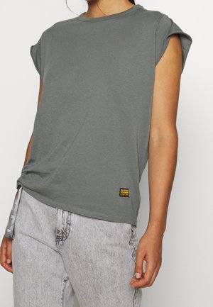 Person med en dämpad grön kortärmad shirt med uppvikta ärmar och ljusgrå jeans, som visar delar av överkroppen och armarna.