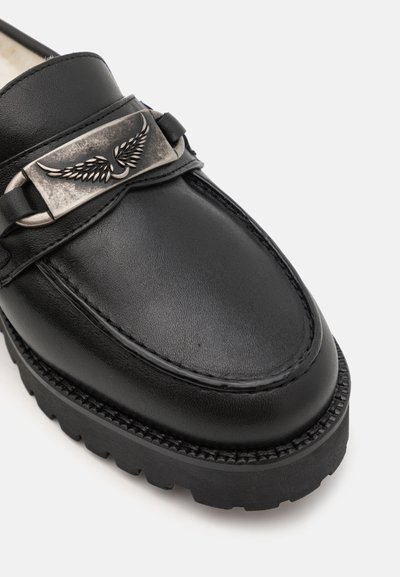 Black leather loafer featuring a silver winged emblem on the strap, textured surface, and a rugged rubber sole with a pronounced tread pattern.