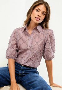 Gennemsigtig mauve-bluse med tekstureret diamantmønster, krave og puffede ærmer. Kombineret med mørke denimjeans.