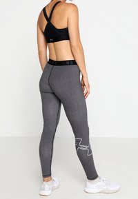 Musta urheiluliivi, jossa on ristikkäiset hihnat, yhdistettynä korkeavyötäröisiin, teksturoituihin harmaisiin legginsseihin, joissa on hienovarainen kuvio ja valkoinen logo.