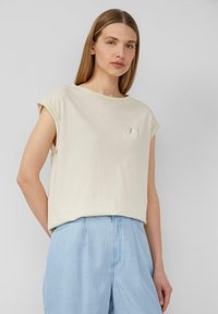 Beige katoenen t-shirt met opgerolde mouwen en een klein geborduurd ontwerp. Gepaarde met lichtblauwe denim broek. Vloeiende textuur.