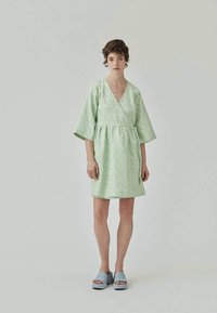 Rochie verde deschis cu decolteu în V și mâneci scurte, evazate. Prezintă o talie strânsă și o siluetă în formă de A. Sandale cu platformă albastre.