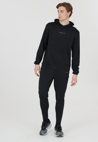 Svart sportig hoodie med texturerad tyg och känguruficka, i kombination med matchande tätsittande joggers och svarta och grå sneakers.