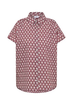 Femme souriante aux longs cheveux bruns portant une chemise à manches courtes à motifs, un pantalon beige large, tenant un grand sac fourre-tout bleu clair, debout contre un mur neutre.