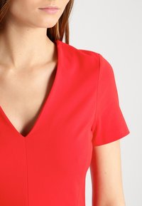Robe rouge à manches courtes avec un décolleté en V, texture lisse et design ajusté, présentant un détail de couture central.