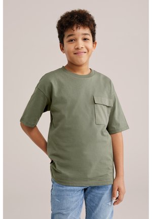Niño con cabello rizado que lleva una camisa de manga corta verde oliva con bolsillo y jeans azul claro, de pie contra un fondo liso beige.