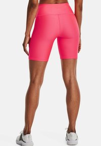 Žena v jasně růžových vysokých sportovních šortkách a bílých teniskách, stojící s nohama od sebe na bílém pozadí.
