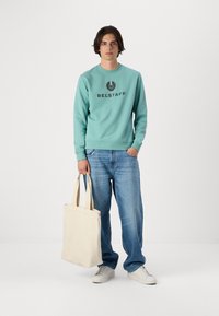 Heled sinine kapuutsiga särk, millel on "BELSTAFF" logo, koos heledate siniste teksade ja valgete tossudega. Hoidab käes beeži kanvasest kott.