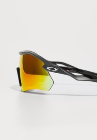 Occhiali da sole sportivi con montatura nera, grandi lenti giallo-verdi e design angolare. Include un logo distintivo e una costruzione leggera.