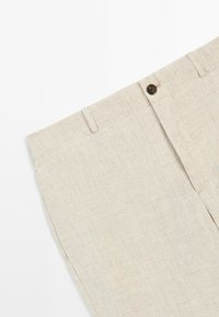 Beige rutig byxa med synlig linning, knapp, bältesöglor och en främre veck på vit bakgrund.