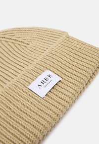 Knit beige beanie with ribbed texture. Features a white label with "ARKK COPENHAGEN" branding. Soft material and foldable design.