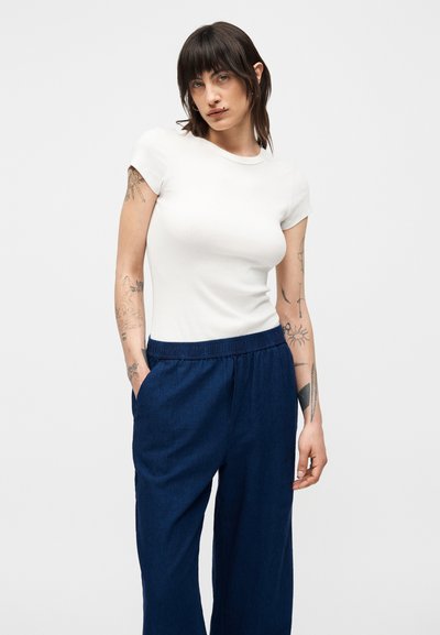 Γυναίκα με σκούρα μαλλιά και τατουάζ φοράει εφαρμοστό άσπρο κοντομάνικο τοπ και φαρδιά σκούρα μπλε παντελόνια με τα χέρια στις τσέπες.