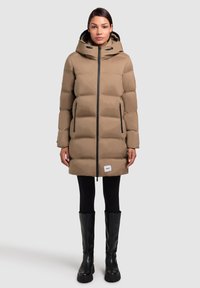 Abrigo marrón acolchado con capucha, cremallera negra, bolsillos laterales y un parche con logo en el dobladillo. Lleva puesto sobre leggings negros y botas altas negras.