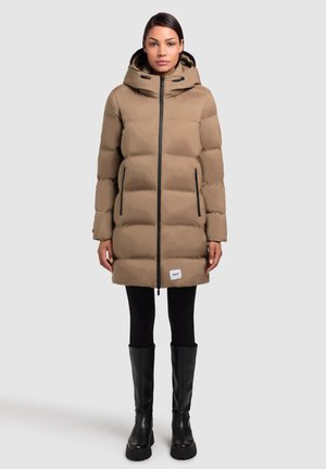 Abrigo marrón acolchado con capucha, cremallera negra, bolsillos laterales y un parche con logo en el dobladillo. Lleva puesto sobre leggings negros y botas altas negras.