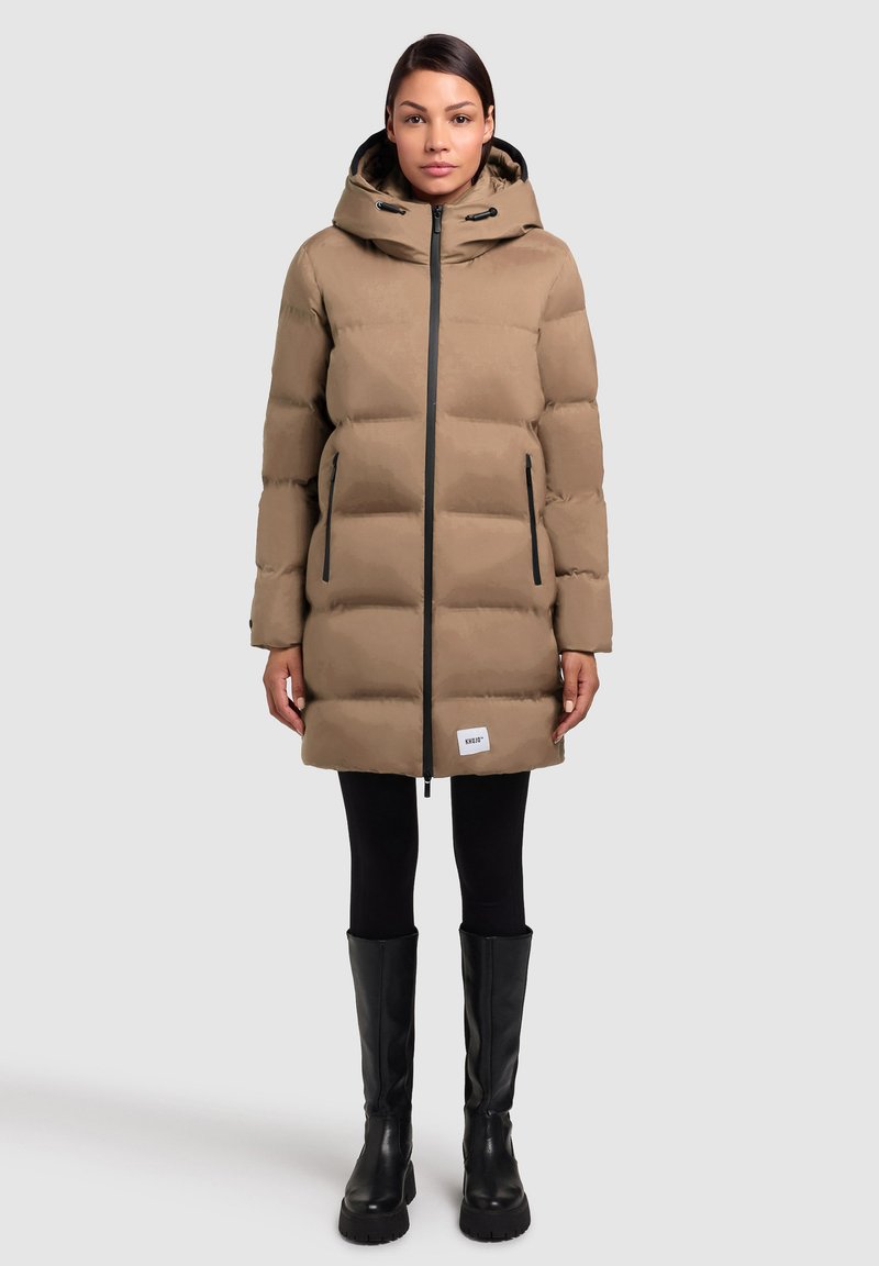 Abrigo marrón acolchado con capucha, cremallera negra, bolsillos laterales y un parche con logo en el dobladillo. Lleva puesto sobre leggings negros y botas altas negras.