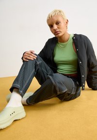 Jachetă bomber neagră, top verde, pantaloni largi cu dungi și saboți menta. Model așezat pe un fundal galben, styling casual.