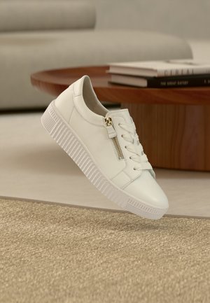 Weißer Ledersneaker mit seitlichem Reißverschluss und Schnürsenkeln, schwebend über einem beigen Teppich, mit einem Holztisch und Büchern im Hintergrund.