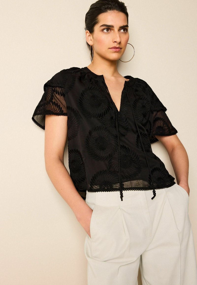 Sort broderet bluse med korte flæser, en V-udskæring og snøre detaljer. Stoffet er gennemsigtigt med blomsterdesigns og bølget kanter.