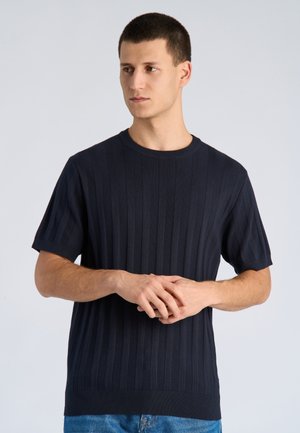 Giovane uomo con capelli corti che indossa una maglietta a maniche corte a coste blu scuro e jeans blu, in piedi con le mani congiunte.