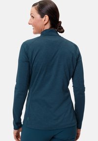 Haut de sport à manches longues en teal avec un col montant, texture lisse et coutures à plat, arborant une petite étiquette verte près de l'ourlet.