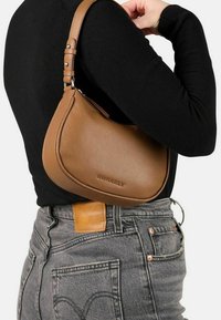 Donna che indossa un top nero e jeans grigi, mentre porta una piccola borsa a tracolla in pelle marrone con tracolla regolabile e nome del marchio impresso.
